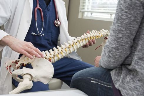 Doctor holding a spine model, explaining it to a patient in an office.