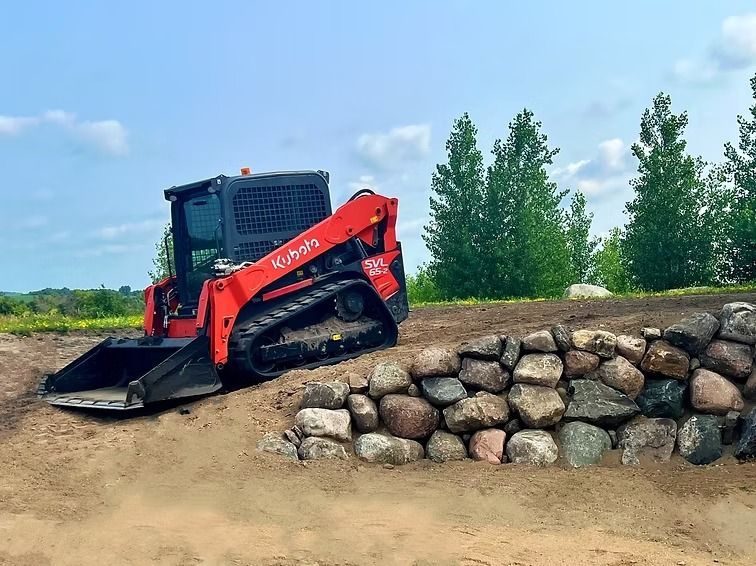 Red Kubota skid steer loader near a stone retaining wall on a dirt slope.
