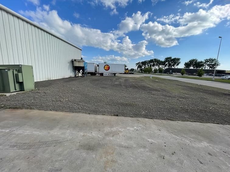 A gravel lot next to a white warehouse with a semi-truck parked behind it, under a cloudy sky.