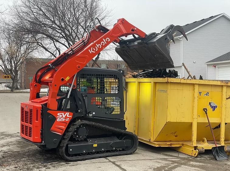 Orange Kubota track loader dumping debris into a yellow dumpster outdoors.
