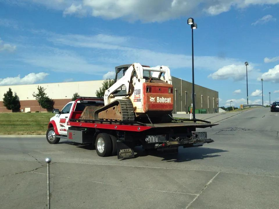 Bobcat skid-steer loader on a flatbed tow truck in a parking lot on a sunny day.