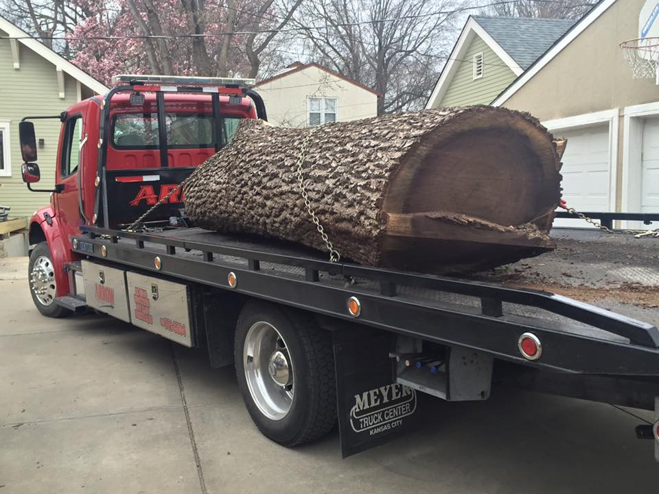 Red tow truck carrying a large tree trunk. The truck is parked in front of a house.