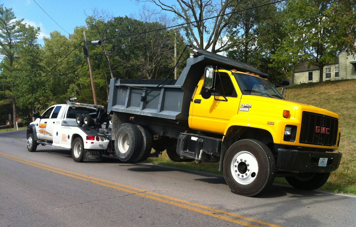 Yellow dump truck being towed by a white tow truck on a road; trees and a building in the background.