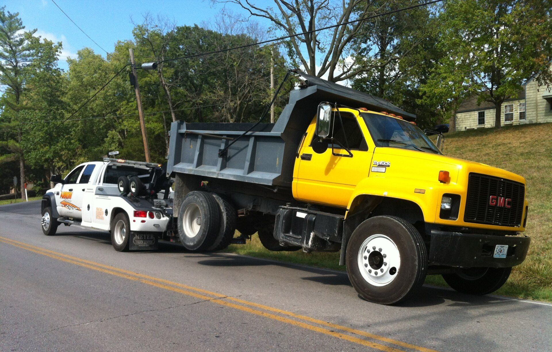 Yellow dump truck being towed by a white tow truck on a road; trees and a building in the background.