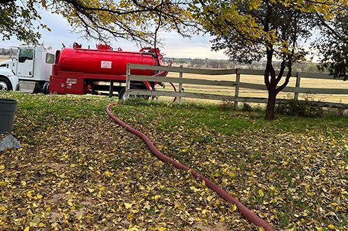A red vacuum truck is parked in a field next to a wooden fence.