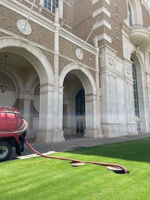 A red hose is connected to a hole in the grass in front of a large building.