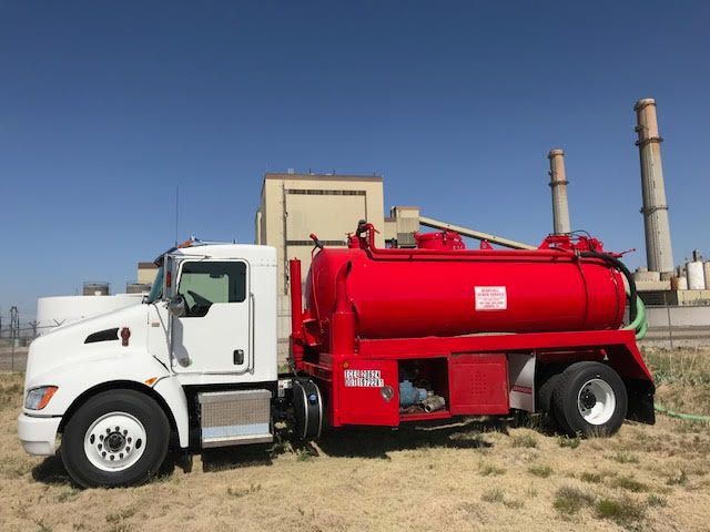 A red and white vacuum truck is parked in a field in front of a factory.
