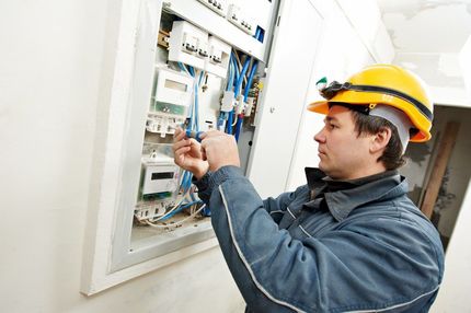 Electrician in a yellow hard hat and blue work jacket works on an open electrical circuit panel on a white wall.