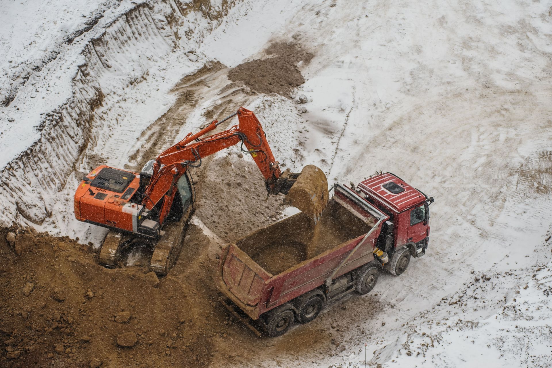 An orange excavator loading soil into a red dump truck on a snow-covered construction site.