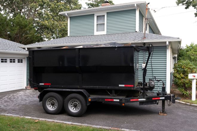 A black dump trailer parked in a residential driveway in front of a house with blue siding and a garage.