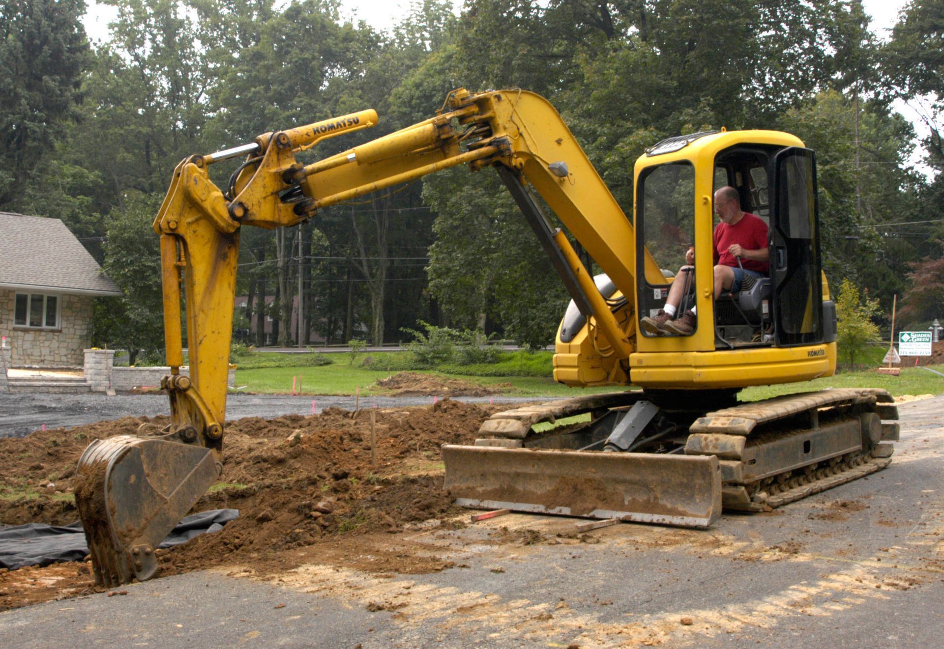 A yellow excavator digging in dirt on an asphalt surface, with a person operating it near a house and trees.