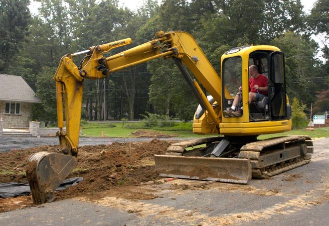 A yellow excavator digging in dirt on an asphalt surface, with a person operating it near a house and trees.