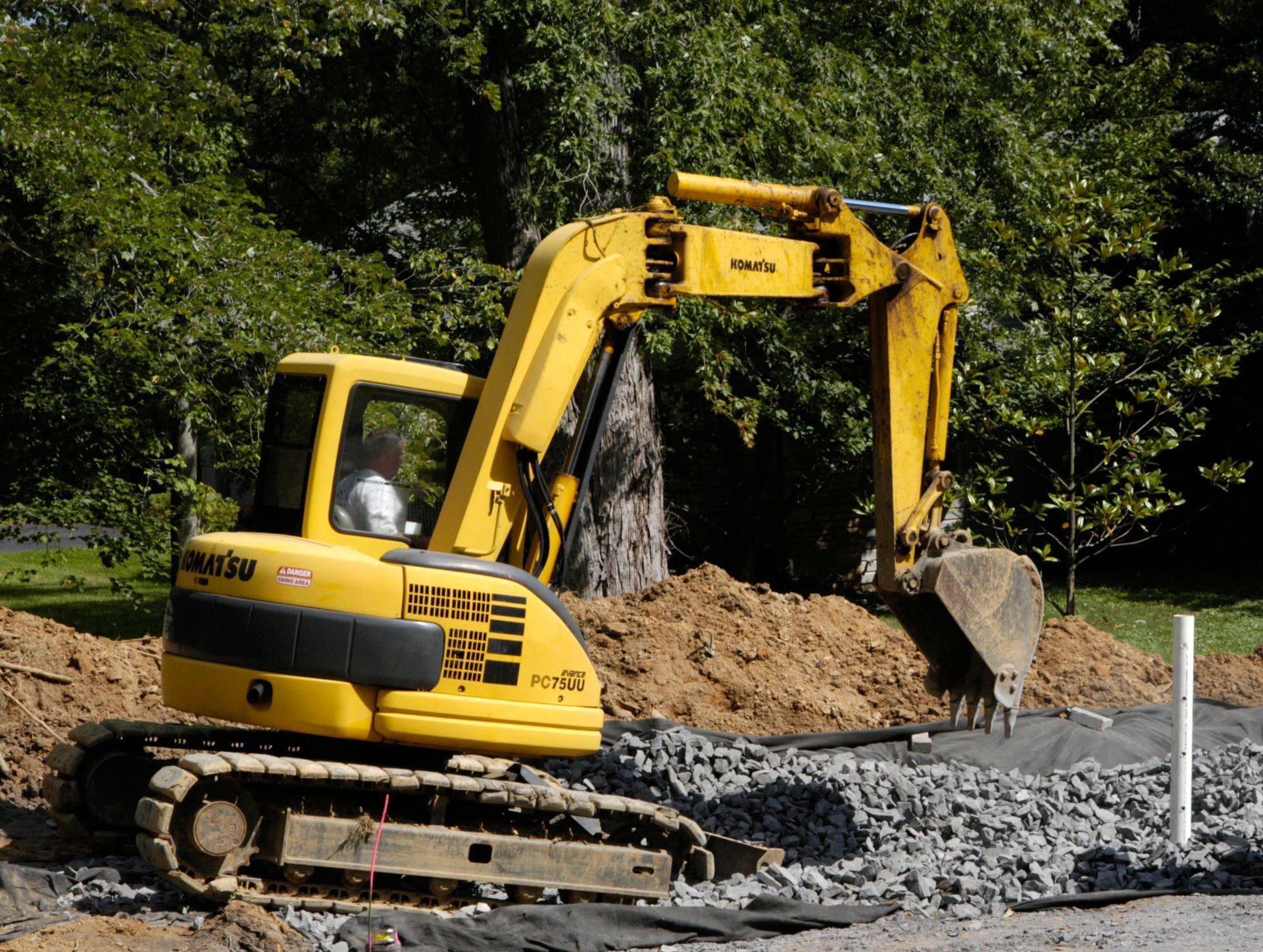 A yellow Komatsu excavator works on a construction site with piles of dirt and gravel in a wooded area.