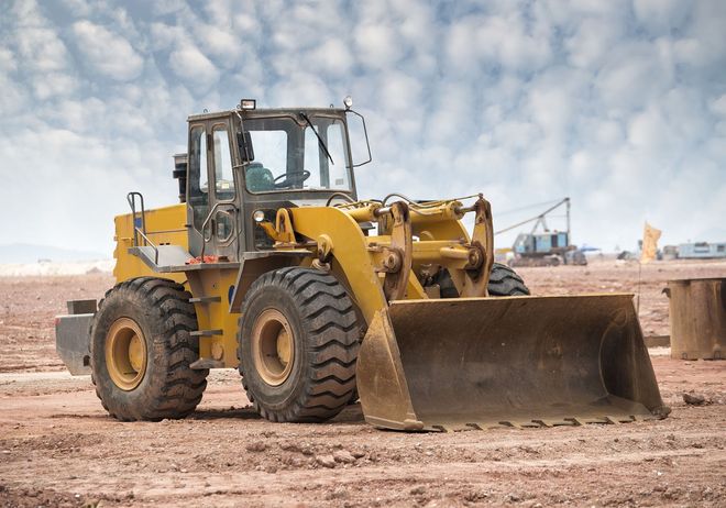 A yellow wheel loader sits on a large, dirt-covered construction site under a cloudy blue sky.