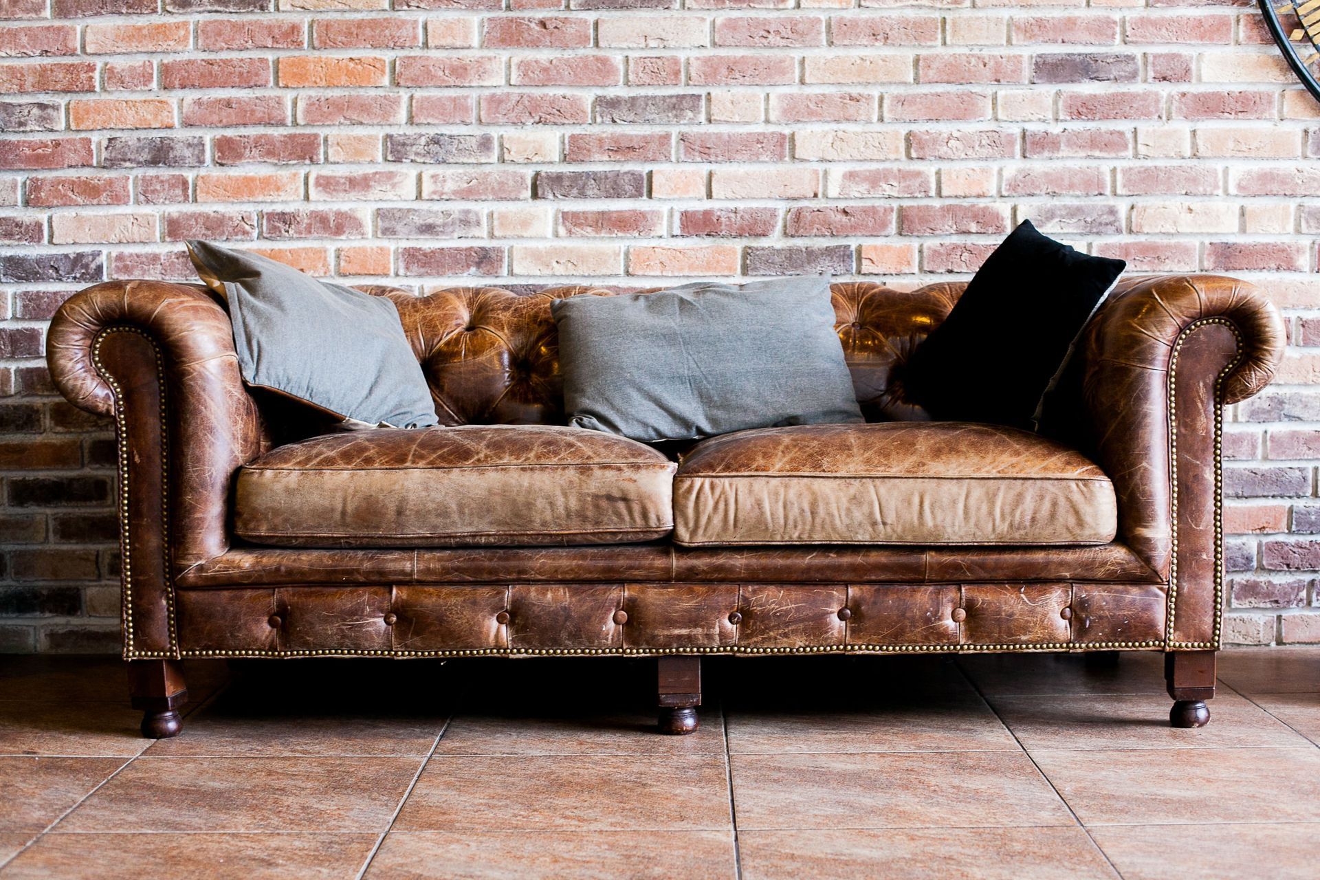 Brown leather sofa with gray and black pillows against a brick wall on a tiled floor