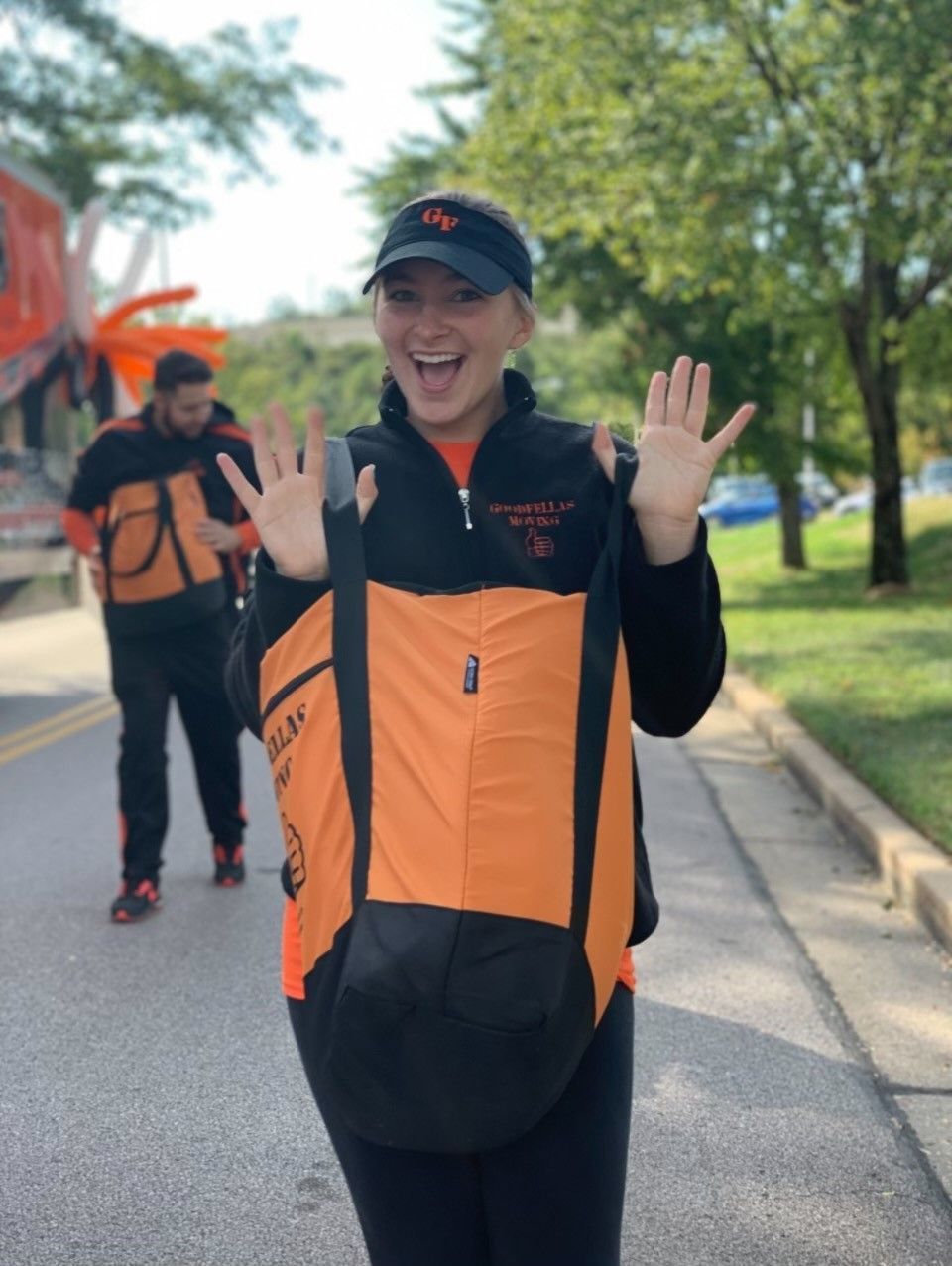 Smiling runner in orange and black raises both hands on a tree-lined road, with another runner behind.