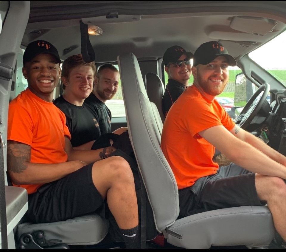 Four people smiling inside a van, two in bright orange shirts, one at the wheel.