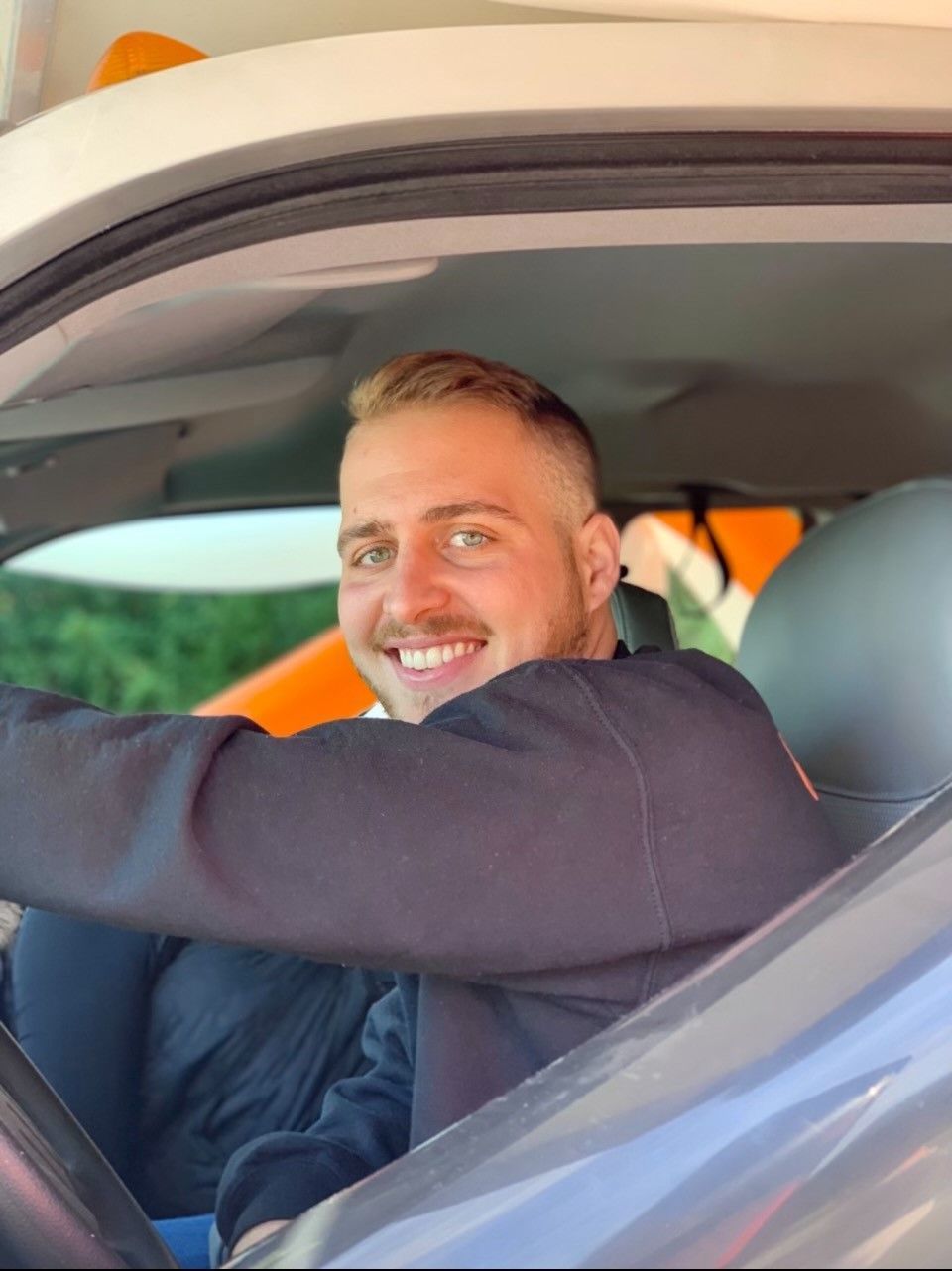 Smiling man in a dark shirt sitting in a car, seen through the open window.