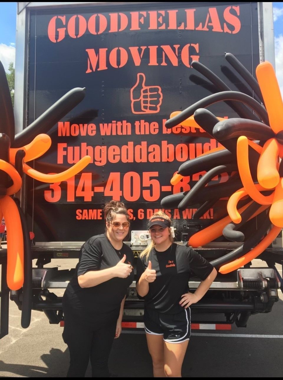 Two people posing in front of a Goodfellas Moving truck with bright orange artwork and phone number.