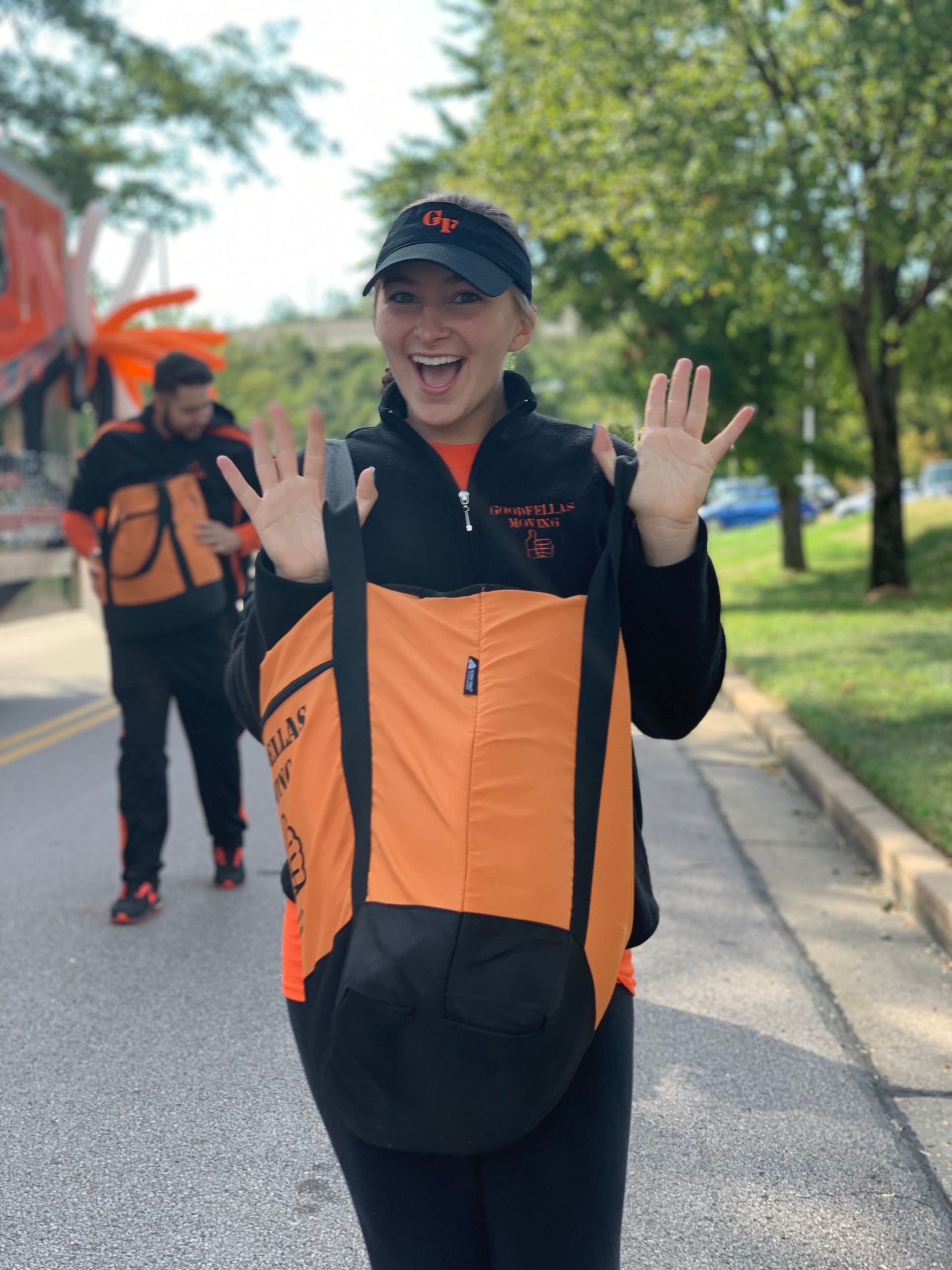 Smiling runner in black and orange gear waving on a tree-lined road, with another runner behind.