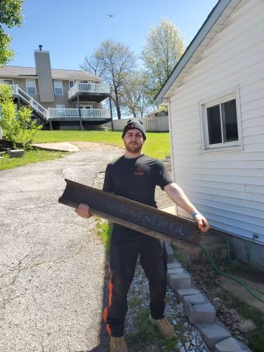 Person in a black shirt and cap holding a long metal gutter beside a white house and driveway.