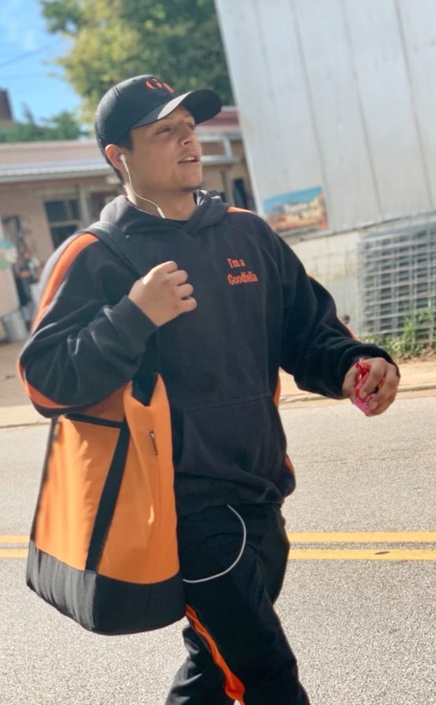 Man walking outdoors in black and orange workwear, holding a cup and a bag, near a building and parked vehicles
