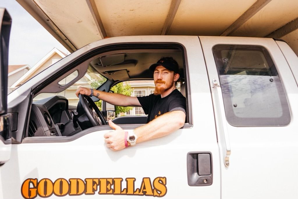 Driver in white truck with “GOODFELLAS” logo, wearing a black cap and holding a drink.
