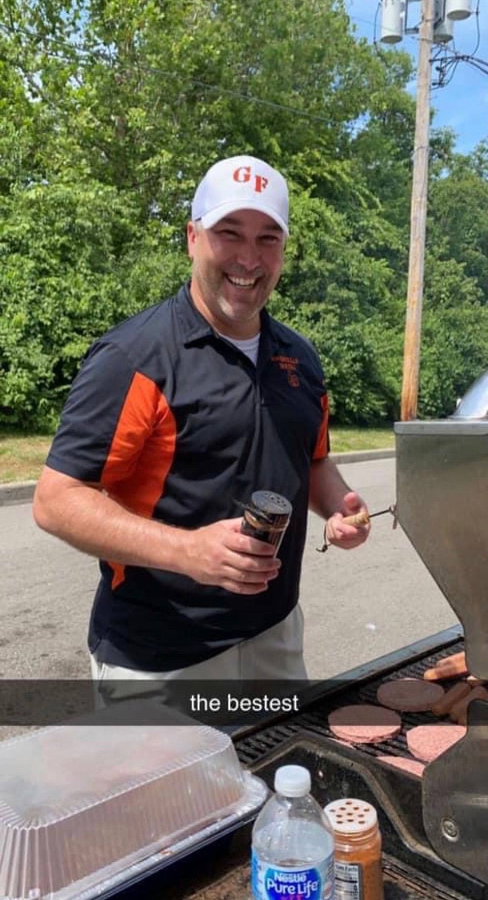 Man smiling beside a grill, holding a drink and utensils outdoors with bottles and grilling tools nearby