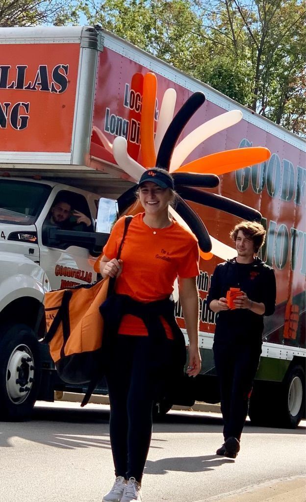 Two people in orange shirts carry large feathered headdresses beside a truck parade float.