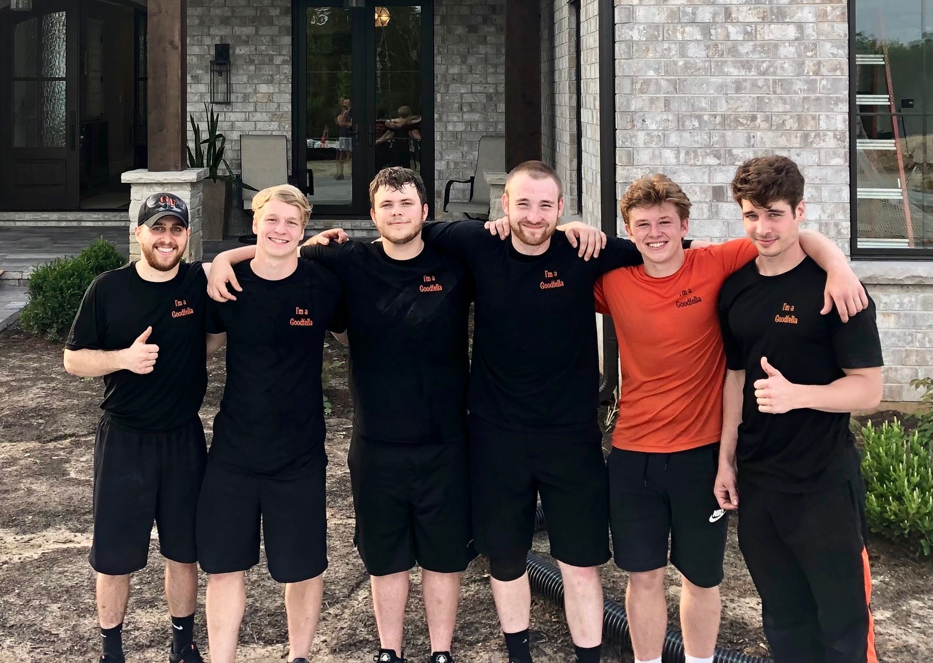 Six teammates posing outdoors, wearing black shirts and shorts, with one in an orange shirt.