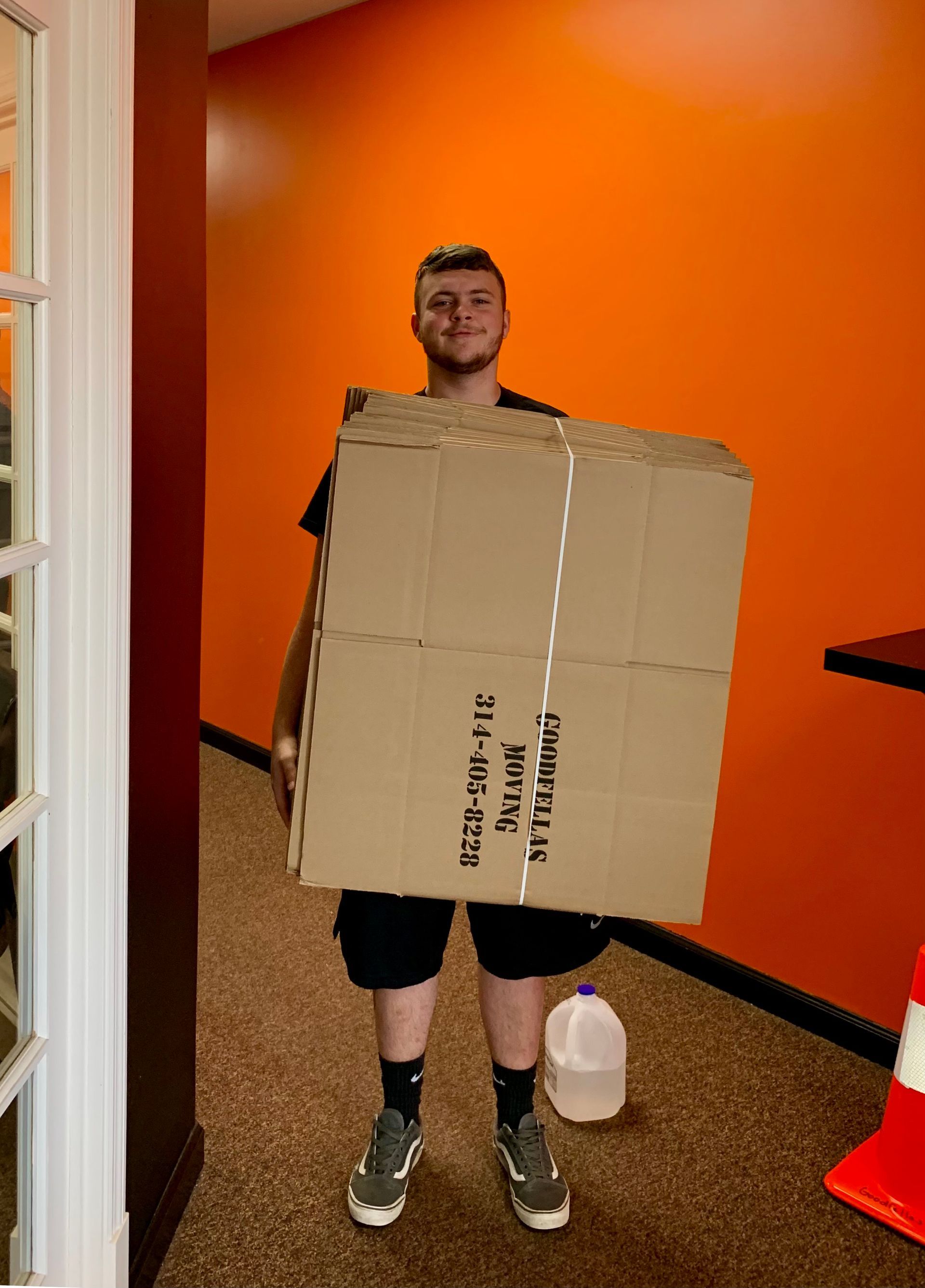Person standing indoors holding a large cardboard box in front of an orange wall, with a jug on the floor.