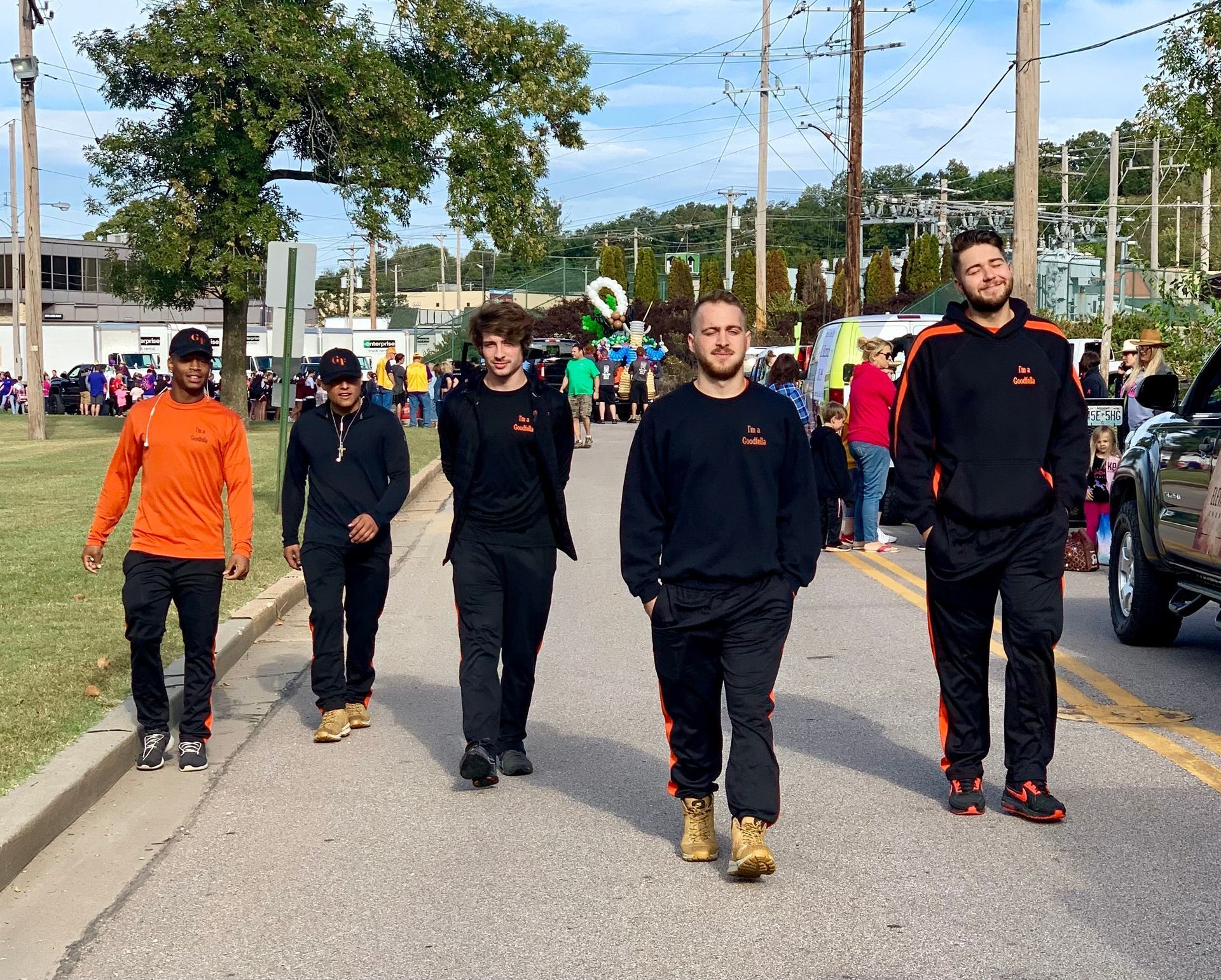 Five people walking down a street in a public park, wearing dark jackets and one orange top.