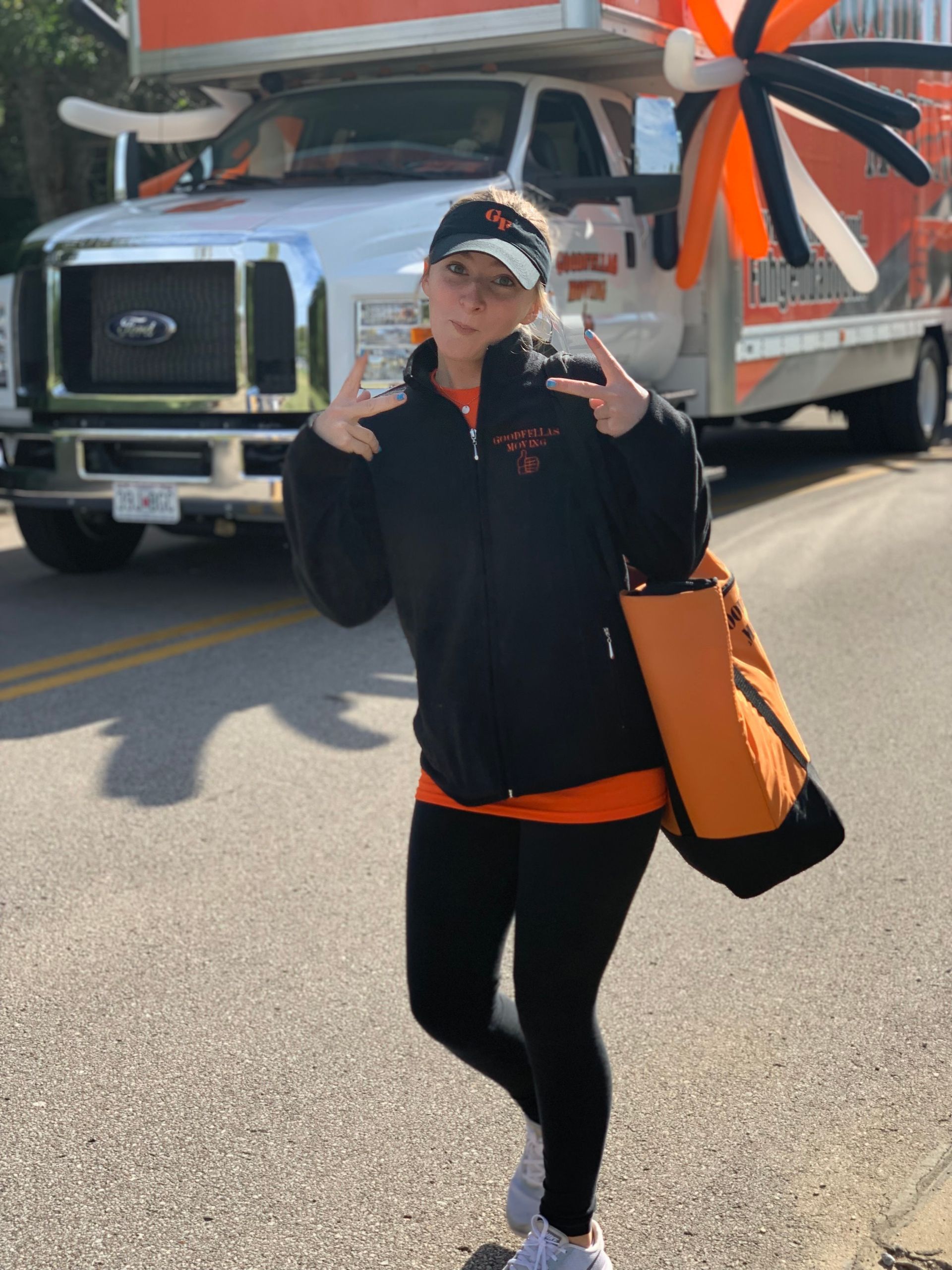 Person posing on a street in front of a truck, wearing black clothes and holding a large orange bag.