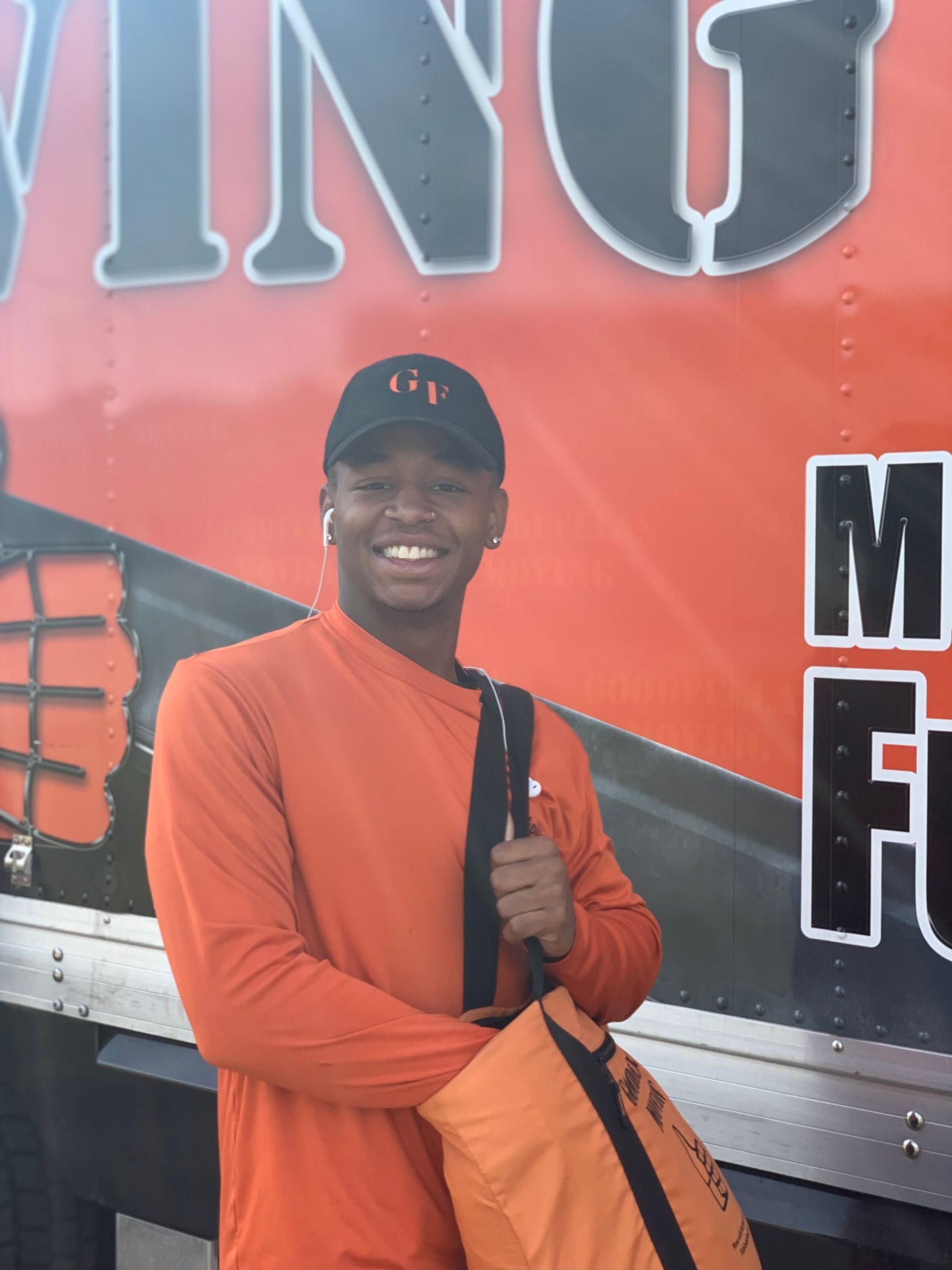 Smiling person in orange shirt and cap holding an orange bag beside a red truck backdrop