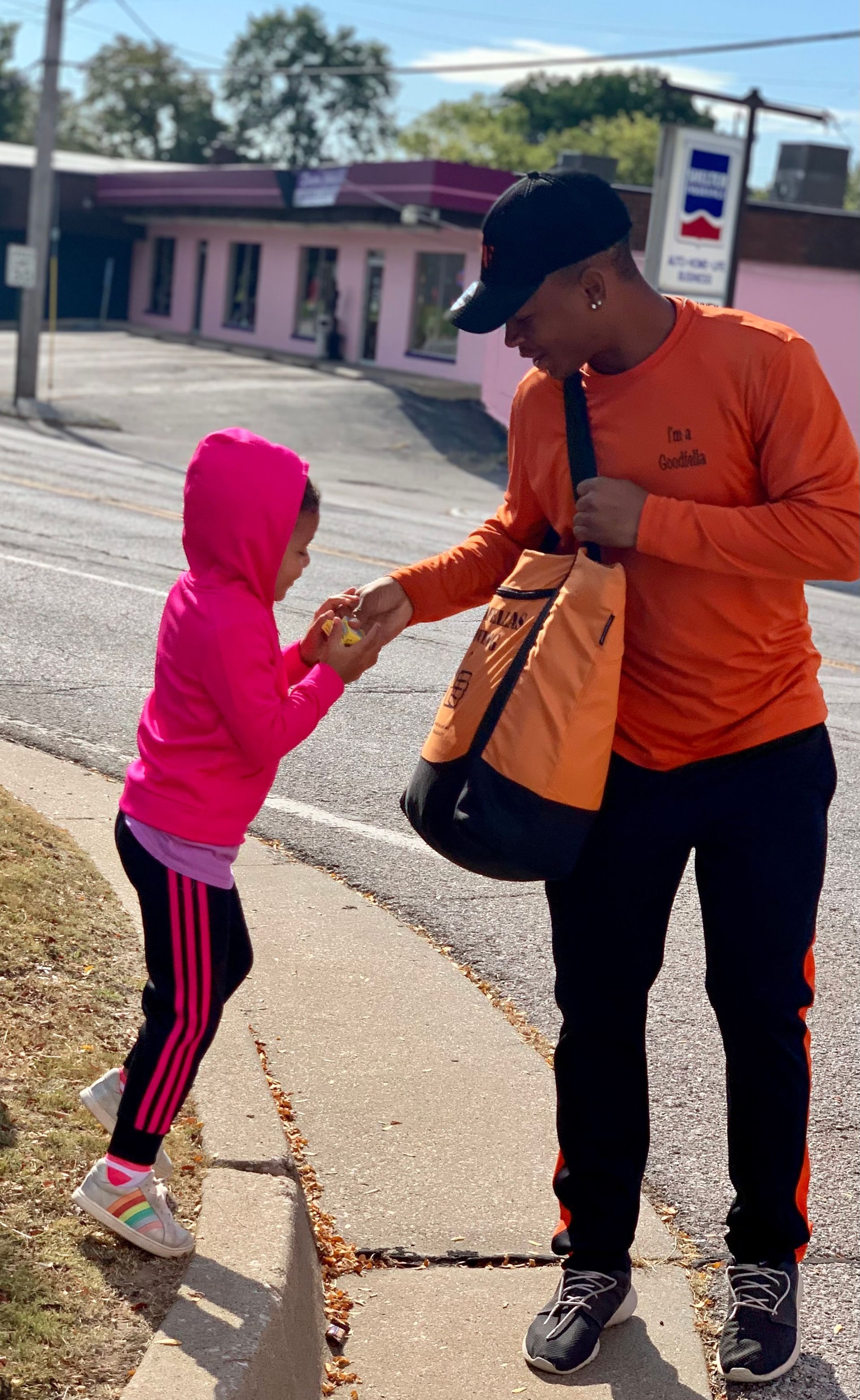 Adult in orange shirt shakes hands with child in pink hoodie on a sidewalk near a road.