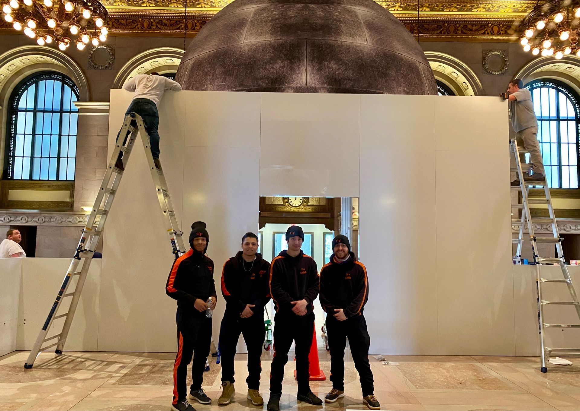 Five people stand in front of a large white display wall with a dome in a grand hall, flanked by ladders.