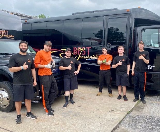Group of staff standing beside a black bus, smiling and giving thumbs-up in a parking lot.