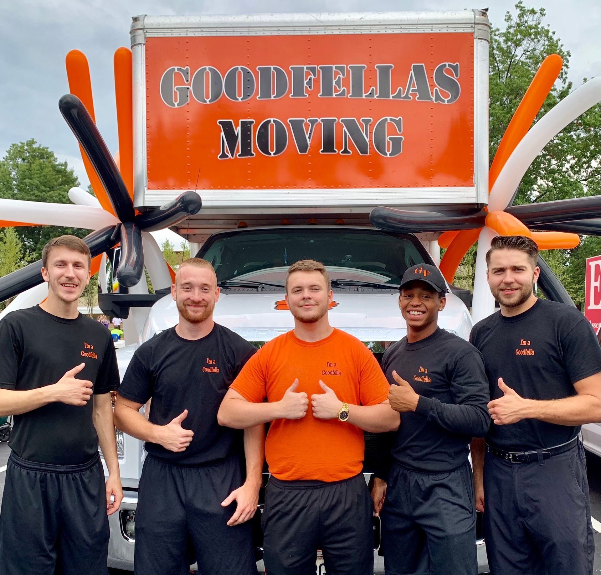 Five movers standing in front of a Goodfellas Moving truck, wearing black and orange uniforms, smiling and thumbs-up.