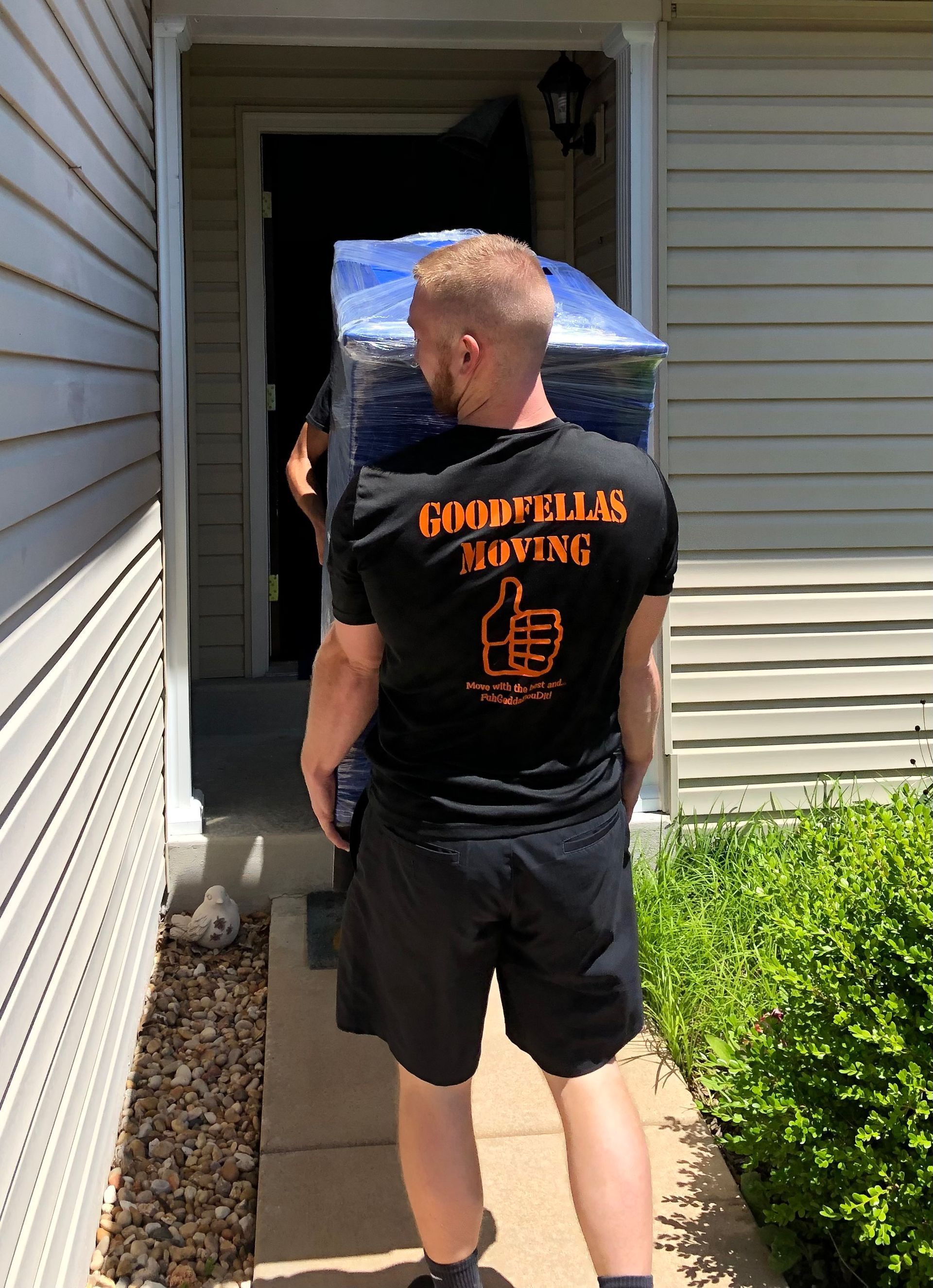Person in black T-shirt and shorts carrying a large box into a house through the doorway.