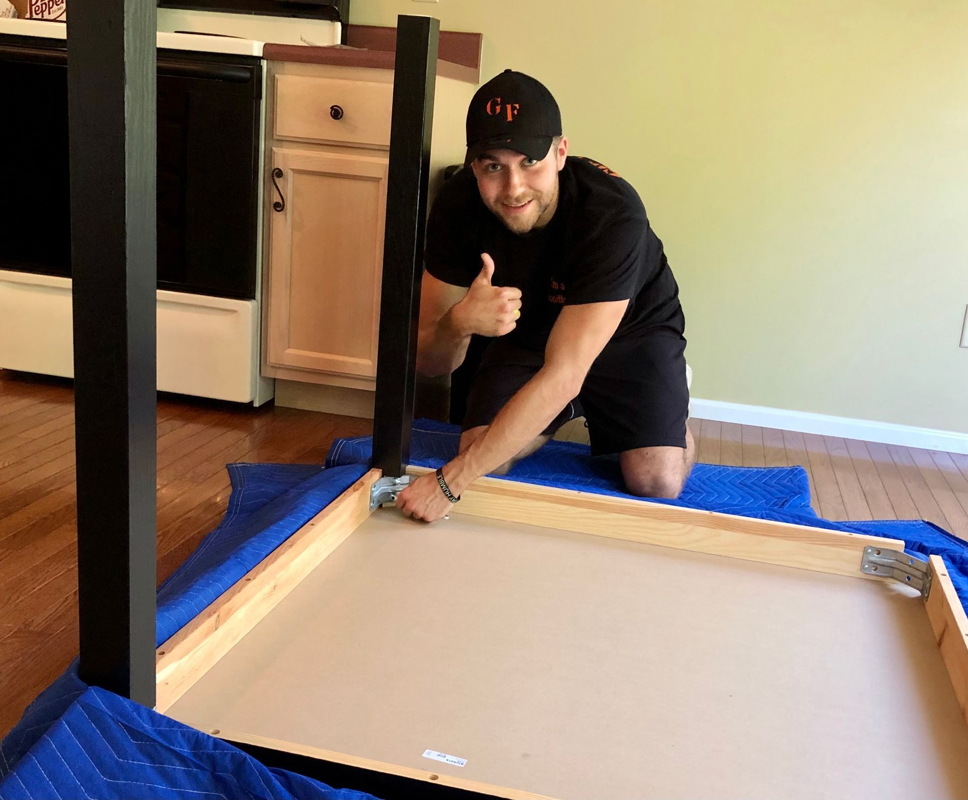 Man kneeling beside an upside-down table, giving a thumbs-up in a room with blue protective padding.