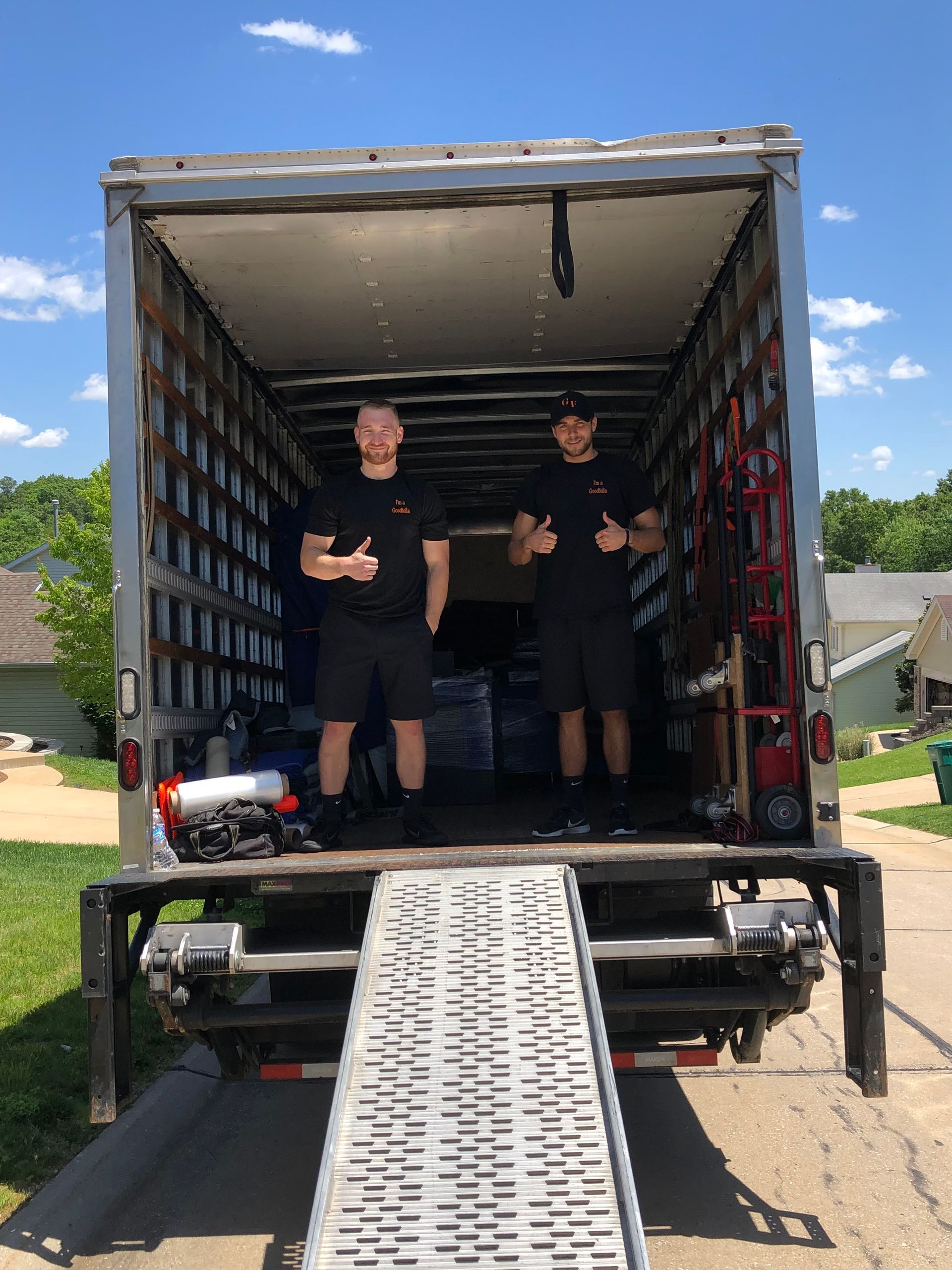 Two workers stand in an open box truck with a metal ramp lowered to the ground.
