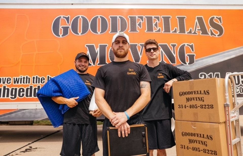 Three movers posing in front of a Goodfellas Moving truck, with cardboard boxes stacked nearby.