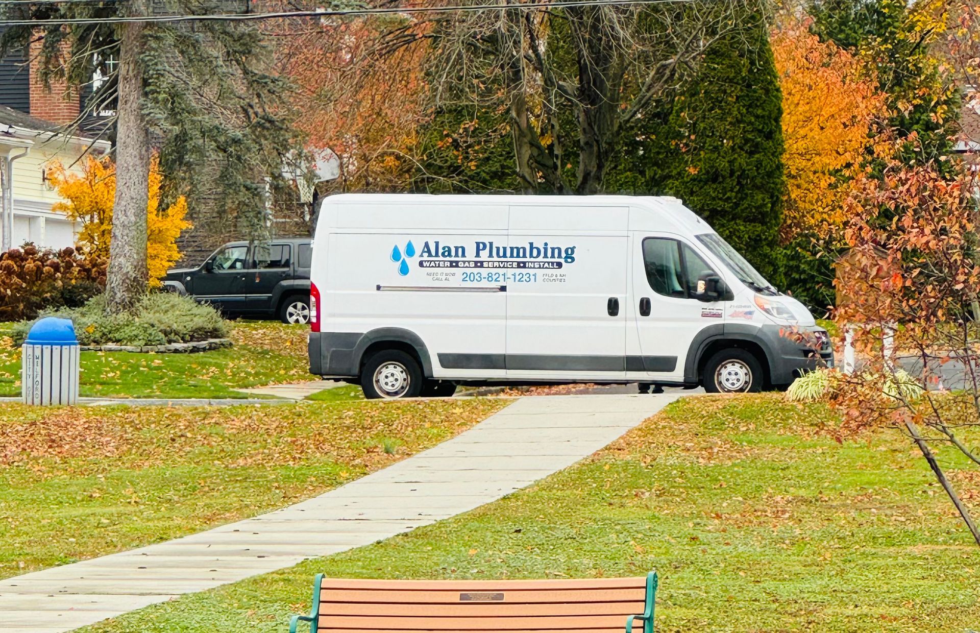 White Alan Plumbing van parked on grass next to a sidewalk and park in autumn.