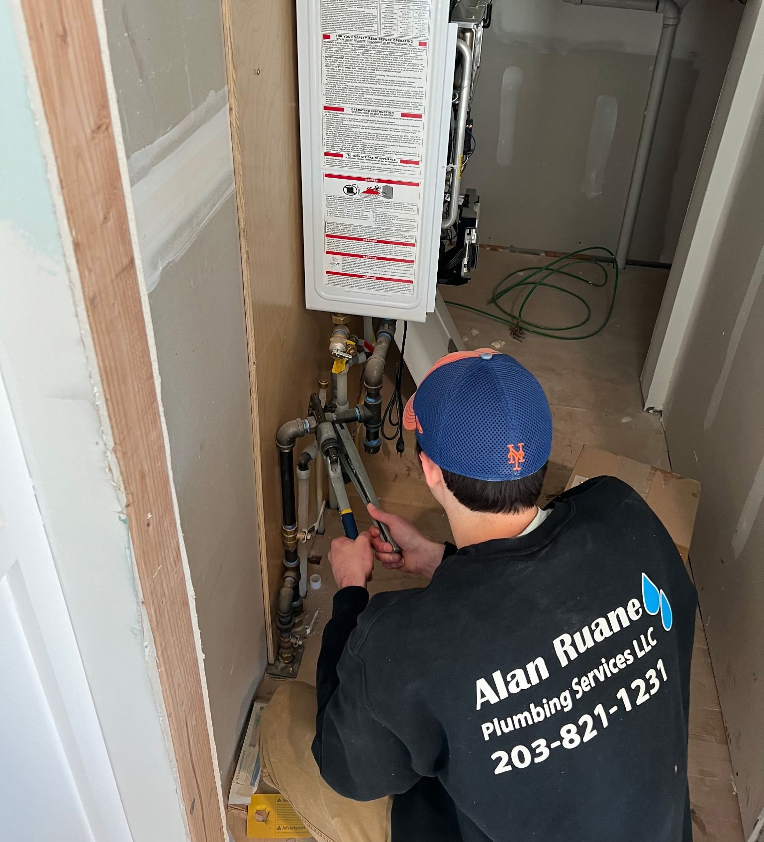 Plumber working on pipes inside a narrow wall space. He wears a blue hat and a black shirt with his company logo.