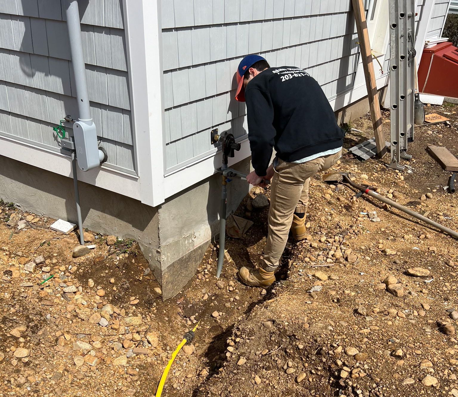 Person using a jackhammer near a building's foundation, excavating dirt. Gray siding, concrete base.