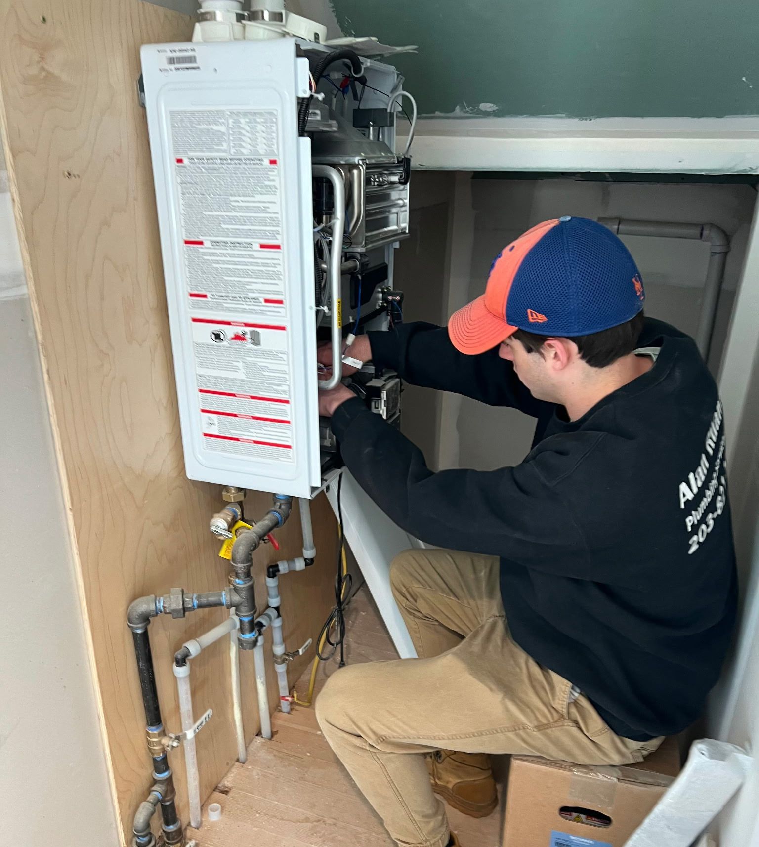 Man in orange hat and black shirt works on a wall-mounted water heater in a narrow space.