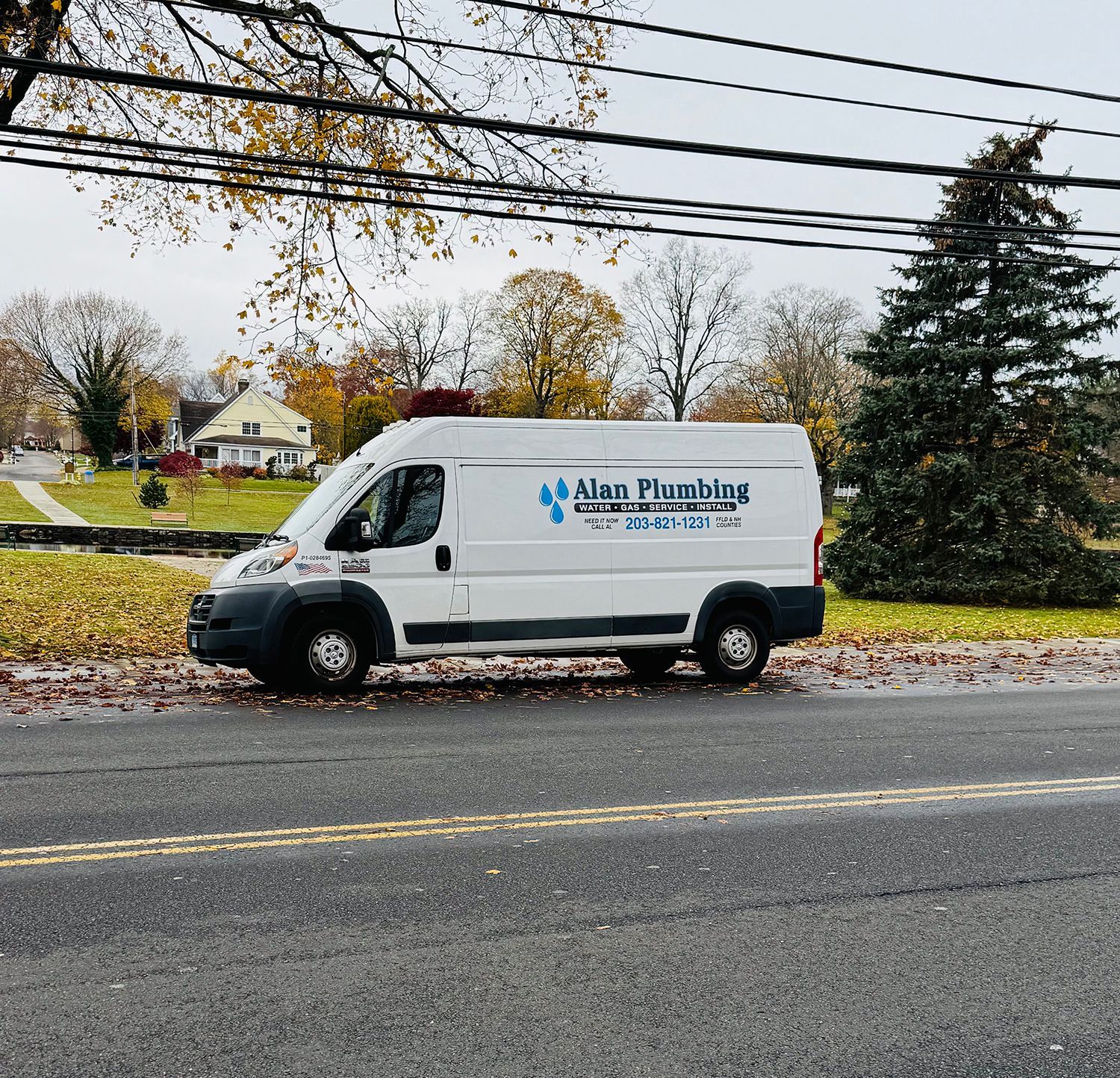 White plumbing service van parked by road, autumn trees in background.