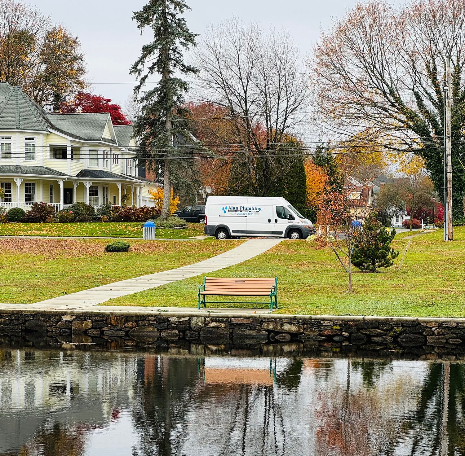 A white van parked near a park bench and pond, with trees and houses in the background.