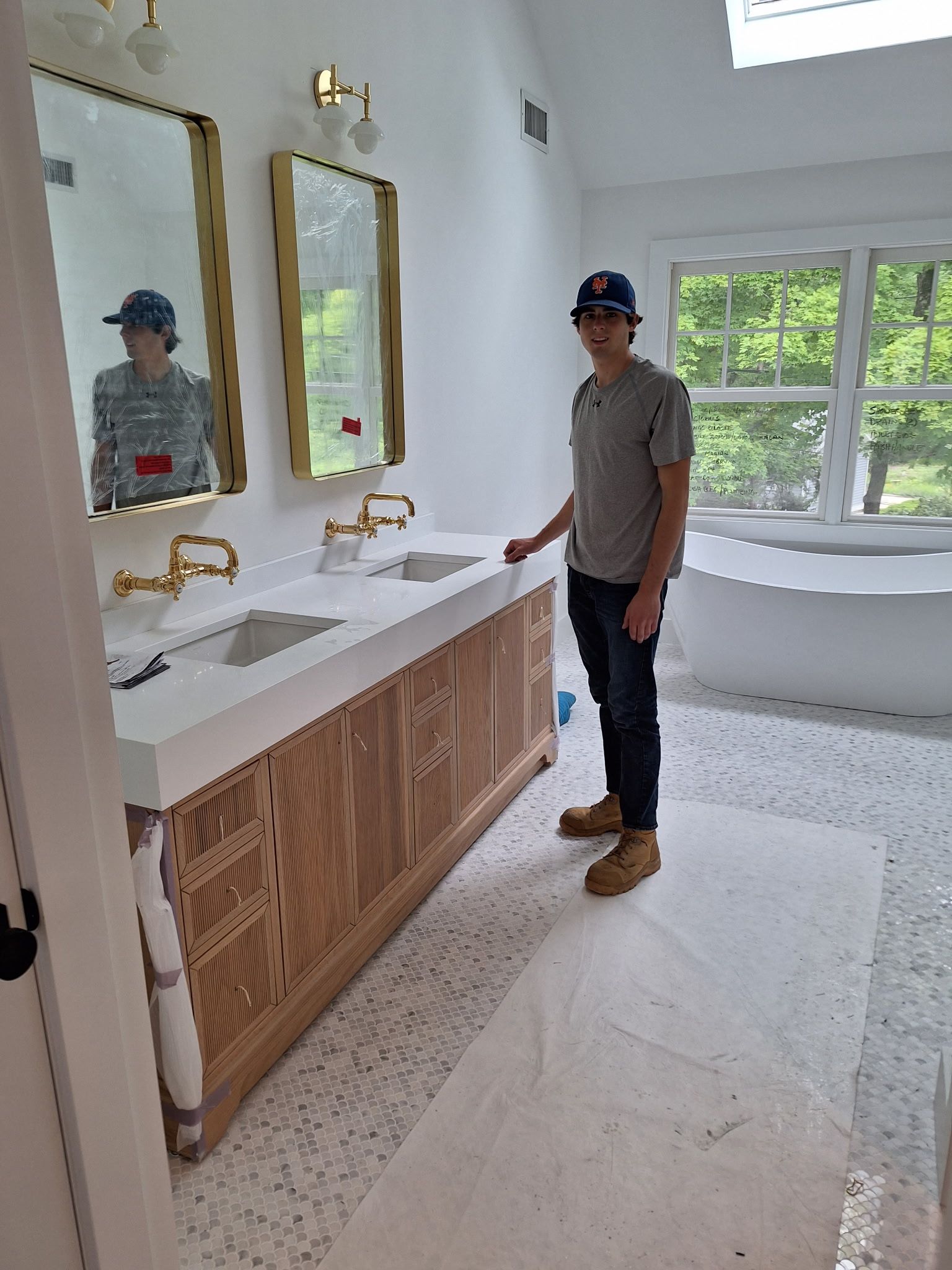 Person stands in a new bathroom with double sinks, mirrors, and gold fixtures. The room has a skylight and a window.