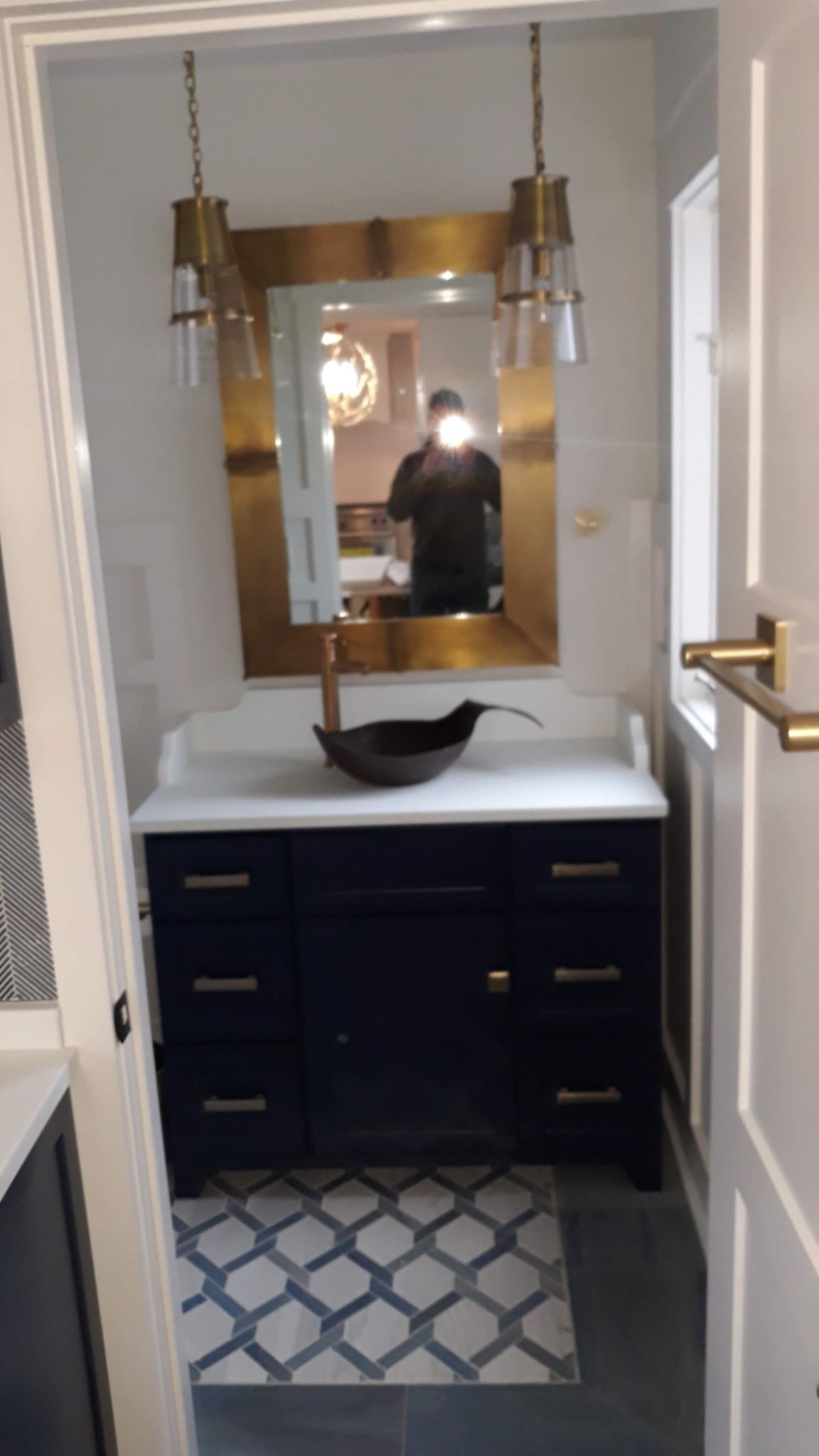 Navy vanity with gold accents, topped with a white countertop and a dark bowl sink, beneath a gold-framed mirror and hanging lights.